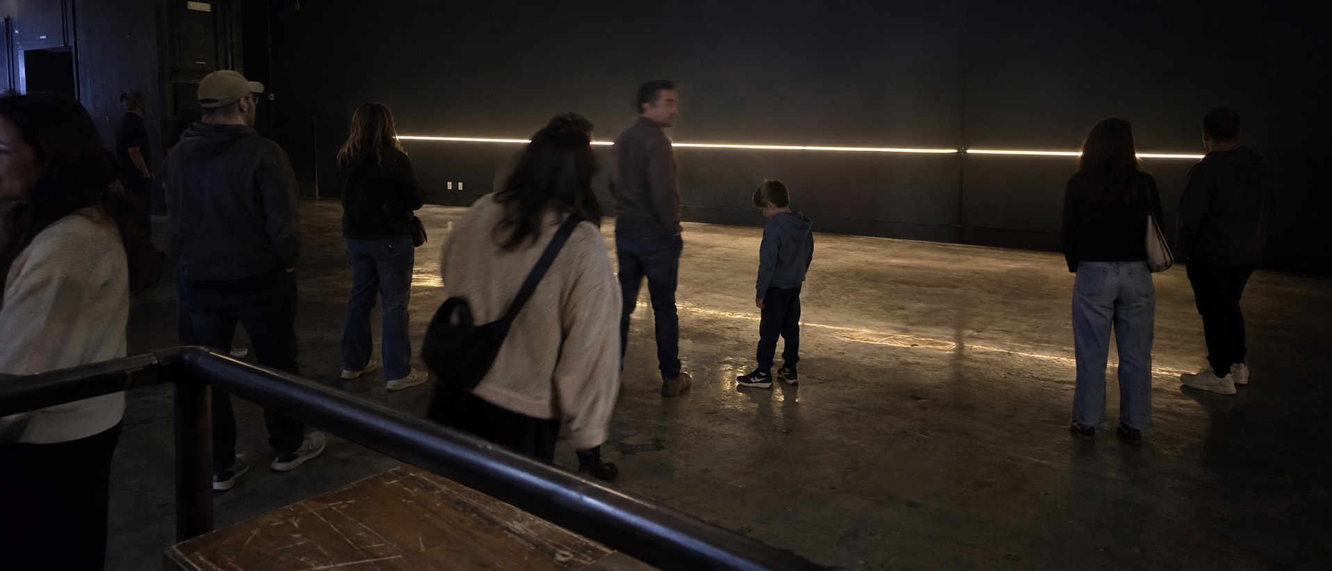A small group of people in a dark room turn toward a thin line of light appearing along the far wall.