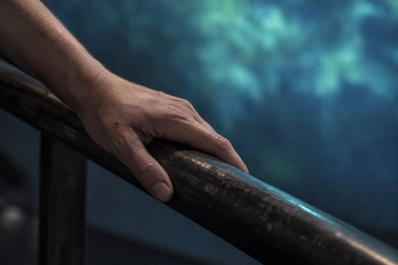 A hand rests lightly on a worn metal rail, sharply lit, in front of a vast blurred field of blue-green projected light.