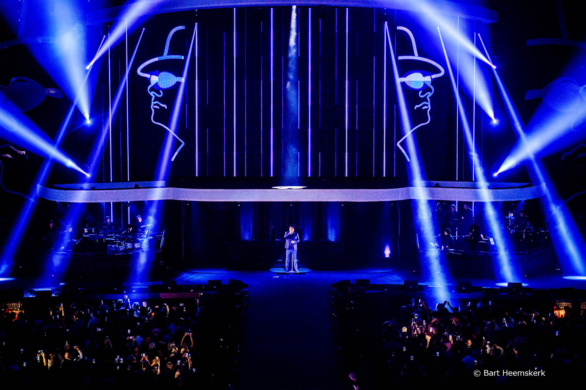 An arena show: line portraits projected across a wide scrim above the stage.