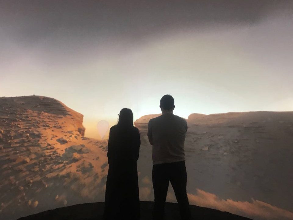 Two visitors standing before a projected desert horizon inside a dome