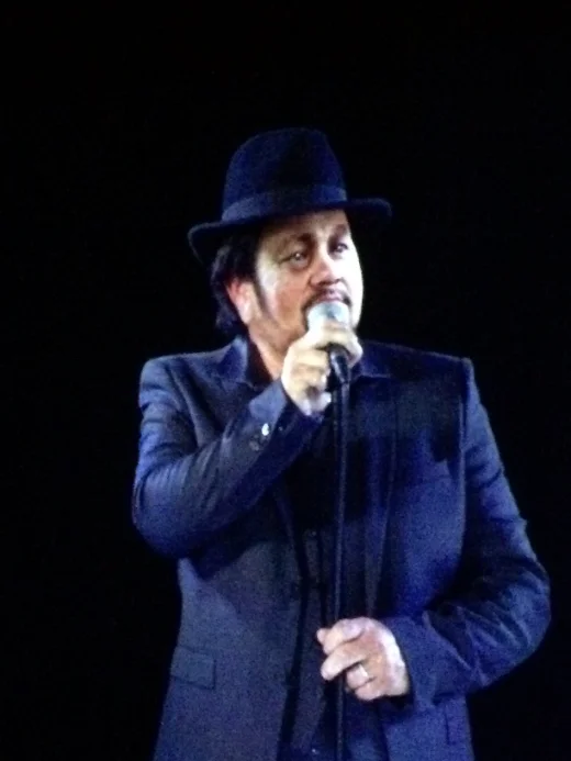 A holographic performer standing in a beam of light on a dark arena stage.
