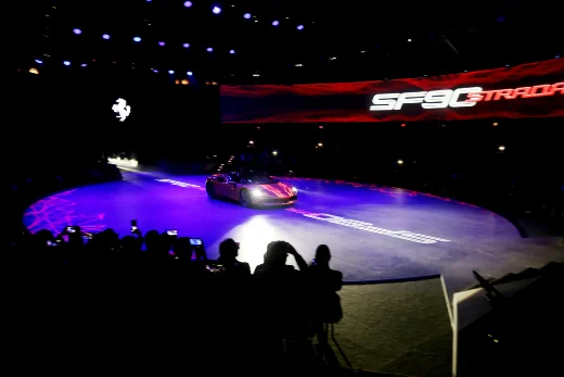 A Ferrari appearing as pure projected light, drawn in moving line work inside a dark theatre in the round.